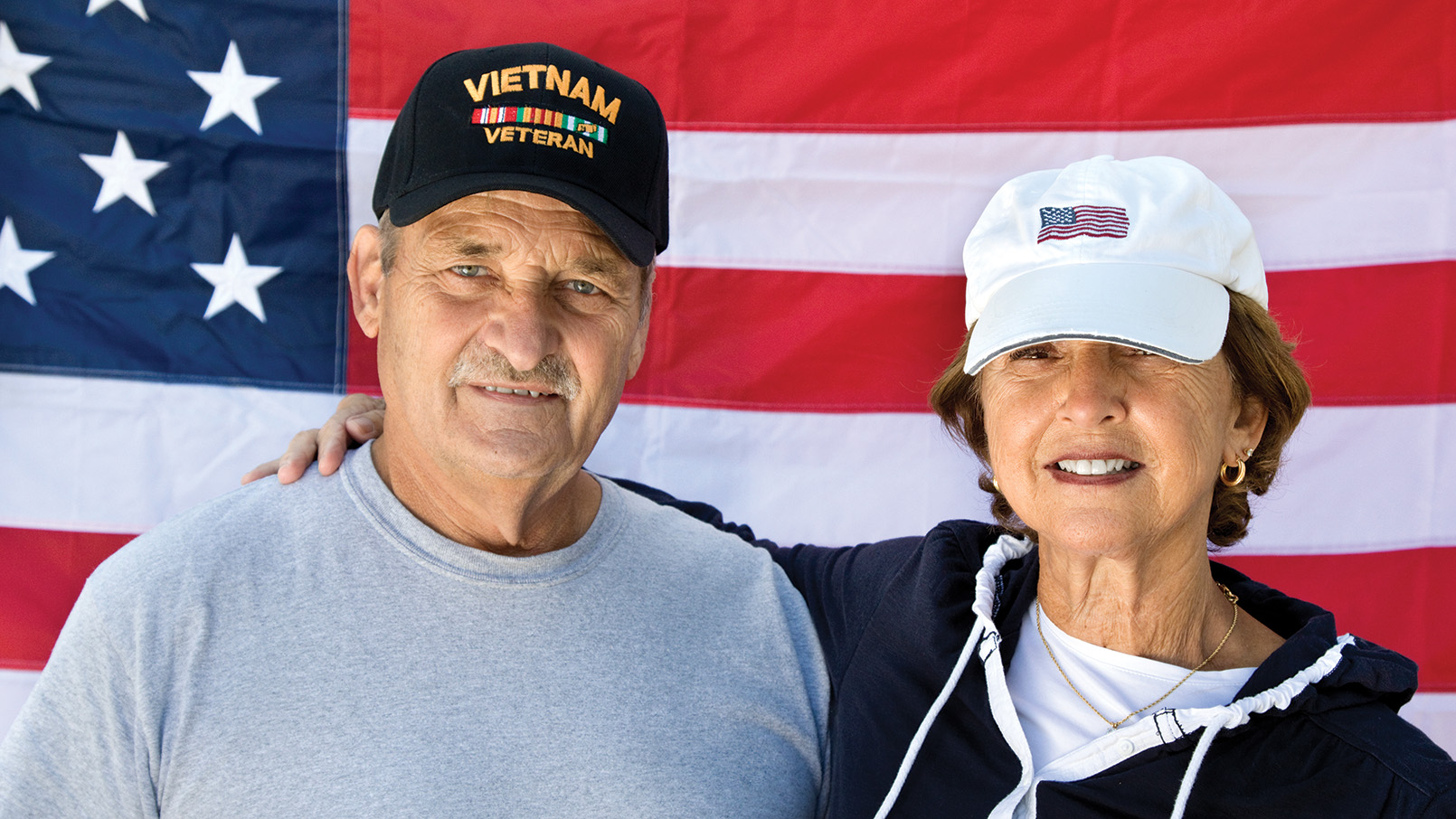 Elderly couple smiling, with the man wearing a Vietnam veterans baseball hat and an American flag in the background