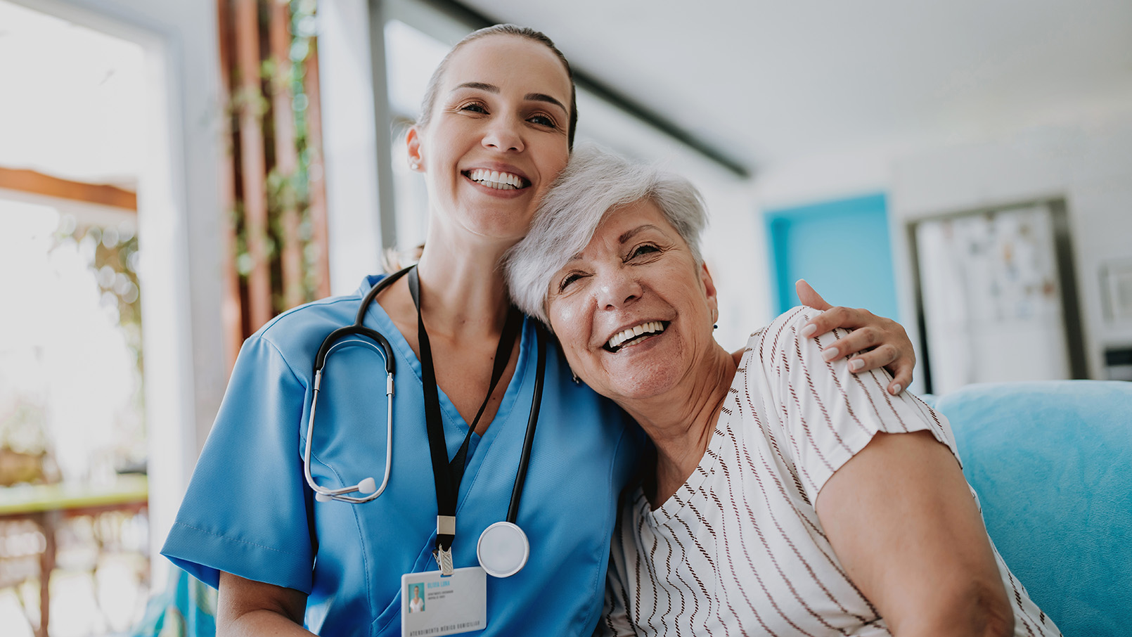 Smiling female nurse in blue scrubs with a stethoscope around her neck embracing a happy elderly woman with short gray hair in a bright home setting.