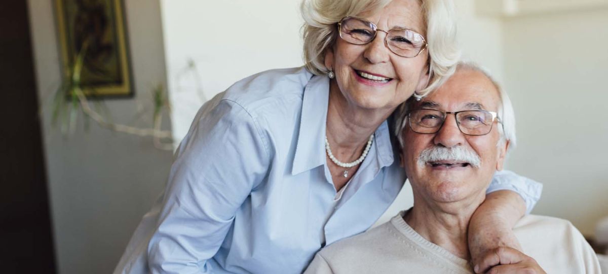 A smiling elderly white couple sit together inside of their Erickson Senior Living apartment home.