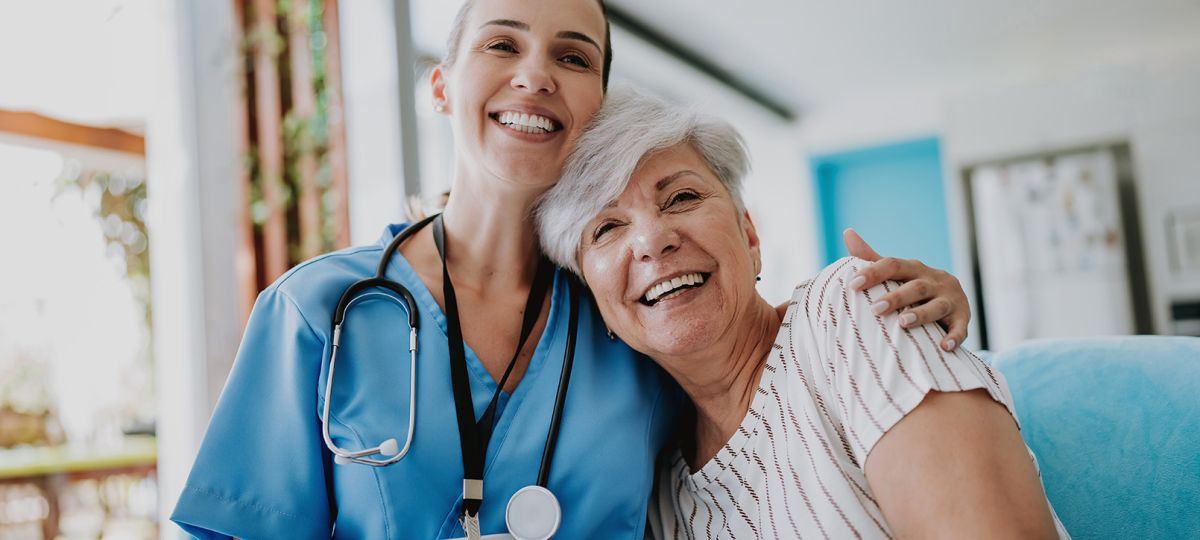 Smiling female nurse in blue scrubs with a stethoscope around her neck embracing a happy elderly woman with short gray hair in a bright home setting.
